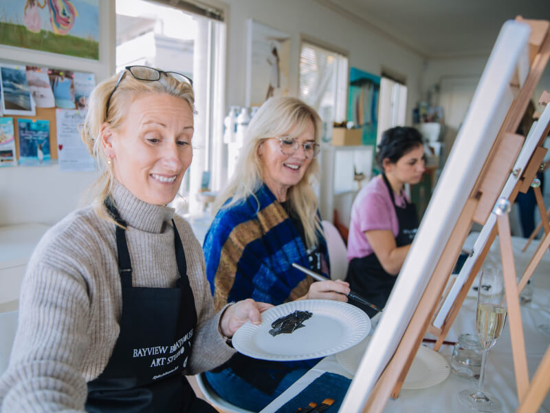 Three women sitting in front of paint canvases smiling.